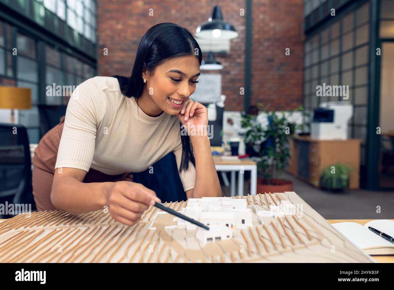 Sorridente biraciale giovane architetto femminile con mano sul mento che analizza il modello di costruzione alla scrivania in ufficio Foto Stock