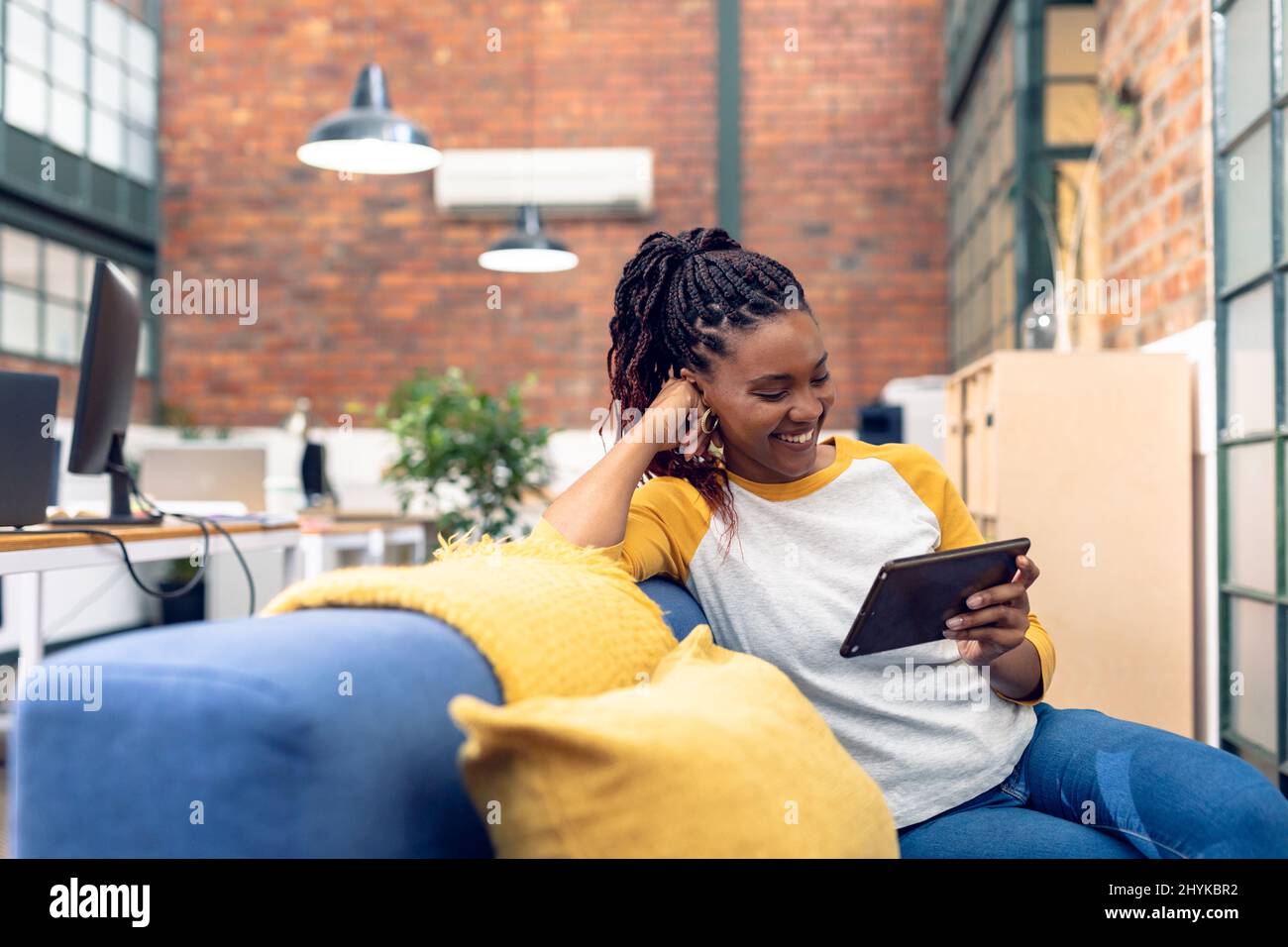 Sorridente giovane donna d'affari afroamericana che tiene un tavolo digitale mentre si siede sul divano in ufficio Foto Stock