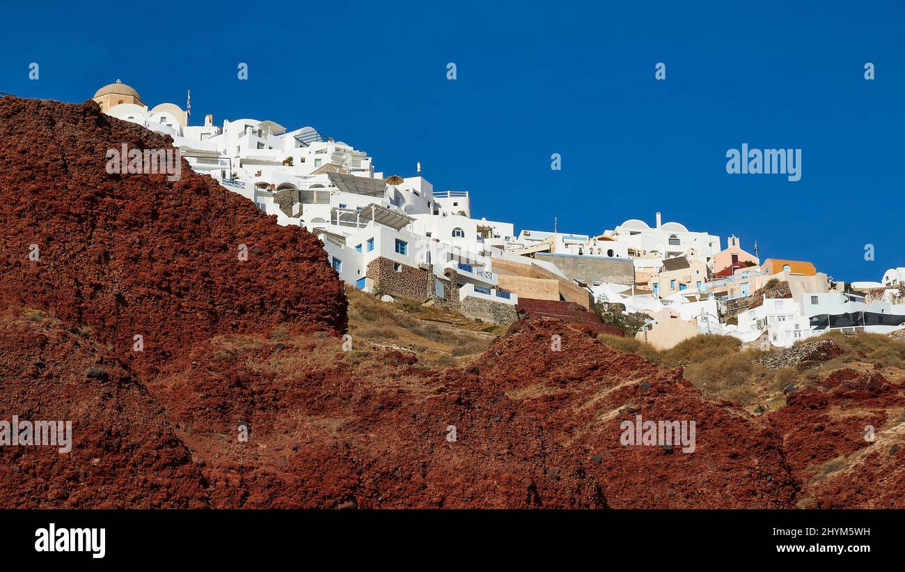 Luce nel pomeriggio, rocce laviche rosse, case bianche e colorate in cima, cielo intensamente blu e nuvoloso, Oia, Santorini Island, Cicladi, Grecia Foto Stock