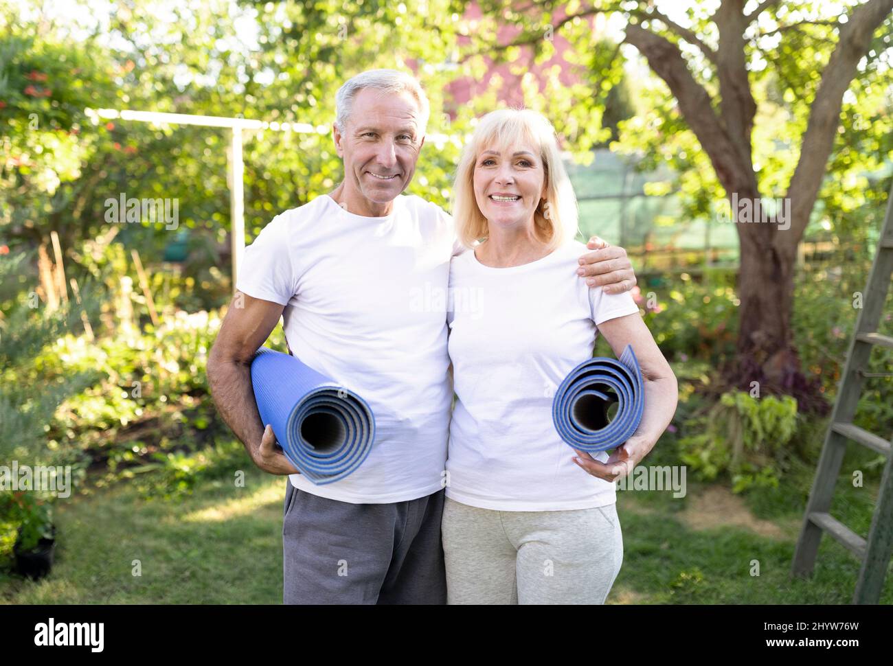 Corpo forte nella vecchiaia. Sposi senior attivi che tengono stuoie yoga e sorridono alla macchina fotografica dopo aver fatto esercizio all'aperto Foto Stock
