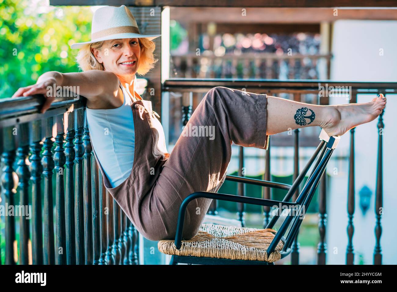 Ritratto di una giovane donna caucasica matura bionda nel suo 50s indossando un cappello e seduto in una posa divertente in una sedia a dondolo sul portico di legno di una cou Foto Stock