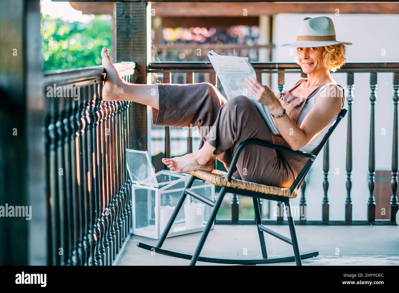 Ritratto di una giovane bionda caucasica matura nel suo 50s indossando un cappello e leggendo un giornale e seduto su una sedia a dondolo in una posa rilassata Foto Stock
