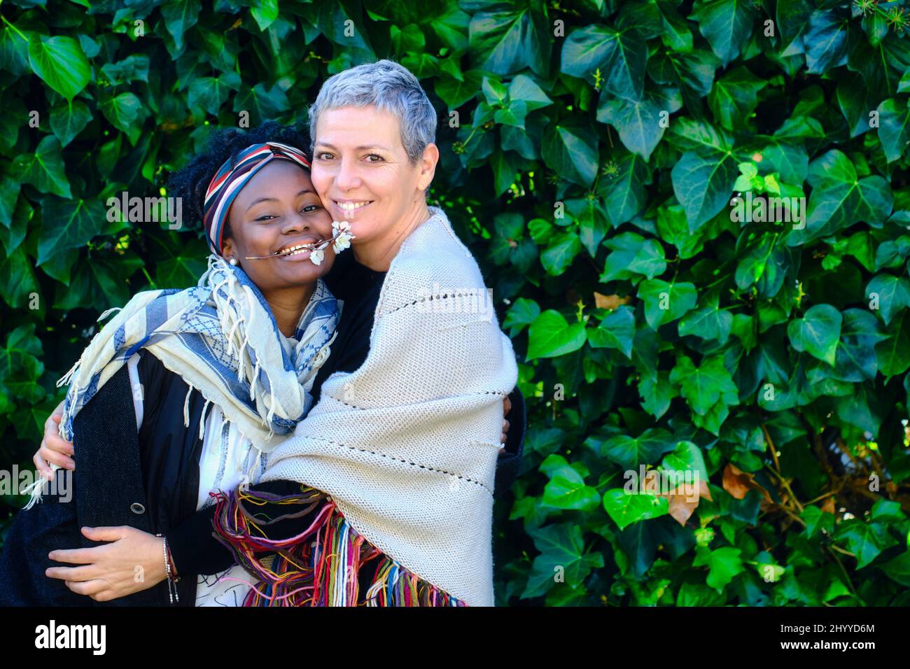 Donna caucasica matura e giovane donna africana che abbracciano in un giardino con un background di foglie. Foto Stock
