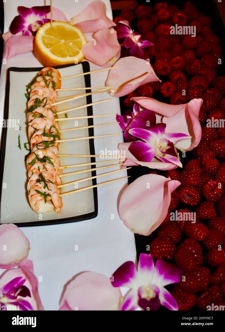 Gamberi su spiedini con menta e zenzero cocktail hors d'oeuvre su vassoio decorato con lamponi, petali rosa e orchidee viola Foto Stock