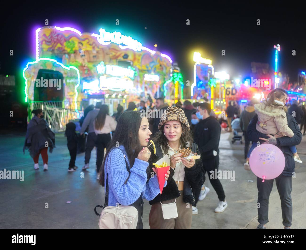 Due donne che mangiano fast food in una fiera notturna affollata e colorata Foto Stock