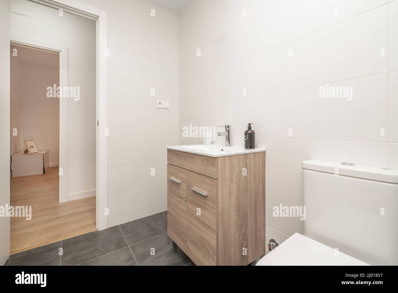 bagno con wc in porcellana bianca, pareti bianche, mobili in legno e pavimenti in gres scuro Foto Stock