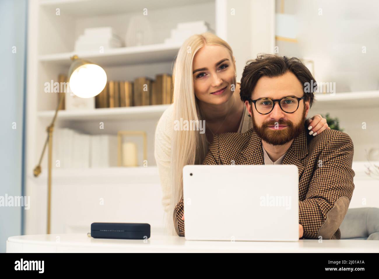 Giovane coppia caucasica sposata ritratto seduto di fronte al portatile sorridendo alla macchina fotografica abbraccio dentro / lavoro remoto concetto . Foto di alta qualità Foto Stock