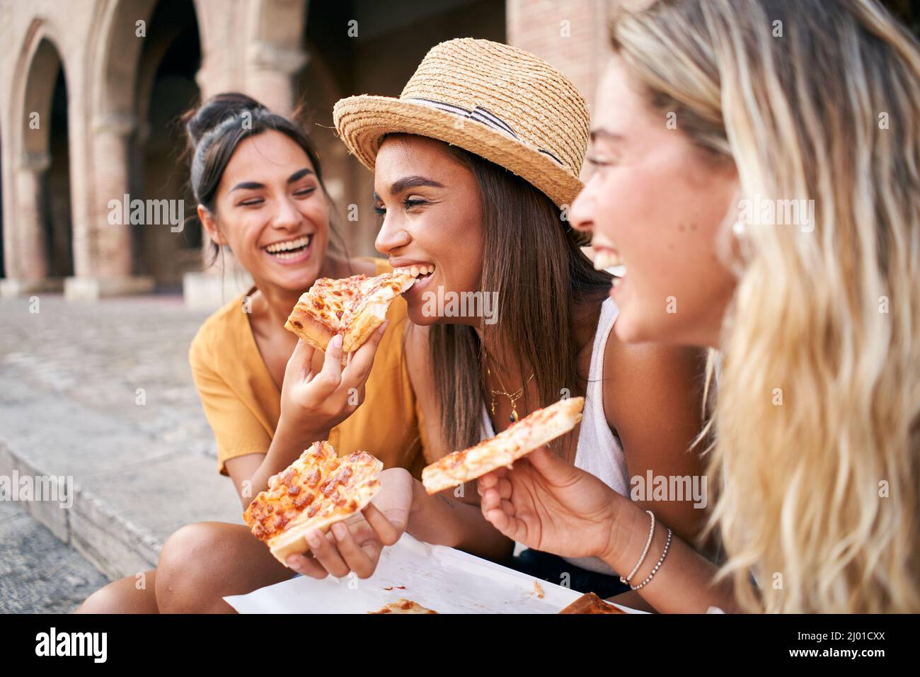Tre belle donne seduti sulle scale delle strade della città mangiare la pizza in una bancarella di strada. Le ragazze felici godono il fine settimana insieme. Foto Stock