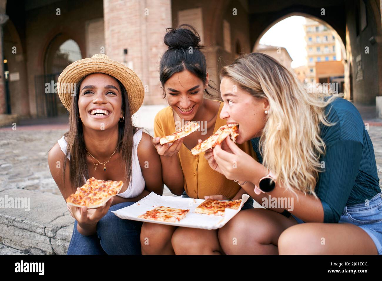 Divertente gruppo misto di tre donne che mangiano cibo di strada all'aperto nella strada della città. Buone amiche che si godono le vacanze estive insieme Foto Stock