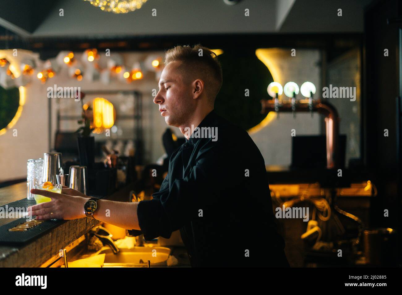 Vista laterale del giovane barman professionista che offre un cocktail pronto al cliente in piedi dietro il bancone del bar sullo sfondo sfocato del moderno nightclub. Foto Stock