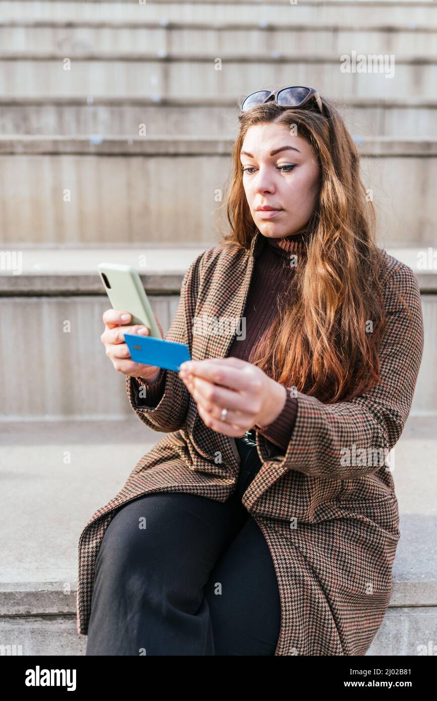 Donna in cappotto a scacchi con capelli lunghi utilizzando carta di credito e telefono cellulare per pagare l'ordine online. Lei è seduta su gradini sulla strada della città Foto Stock
