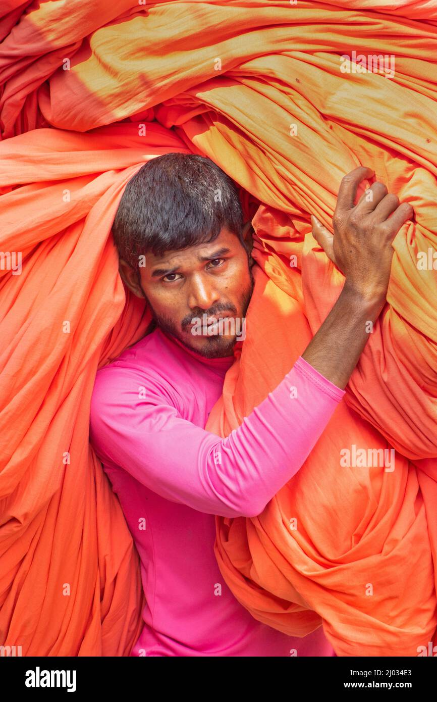 Narayanganj, Dhaka, Bangladesh. 16th Mar 2022. I lavoratori appendono migliaia di diversi tessuti colorati su fili di ferro legati tra un telaio di bambù e costantemente li trasformano in modo che si asciughino perfettamente in campo allagato a Narayanganj, Bangladesh. I fili di ferro sono usati fra una struttura di bambù per generare le linee giganti di lavaggio per la parte finale del processo di tintura mentre i tessuti sono asciugati nel sole. I fili luminosi di panni blu, rosa, arancione e verde-tinti appendono sopra il campo erboso in una rete abbagliante di colori interbloccanti. Questa è la parte finale del processo di tintura dopo la quale il panno è Foto Stock