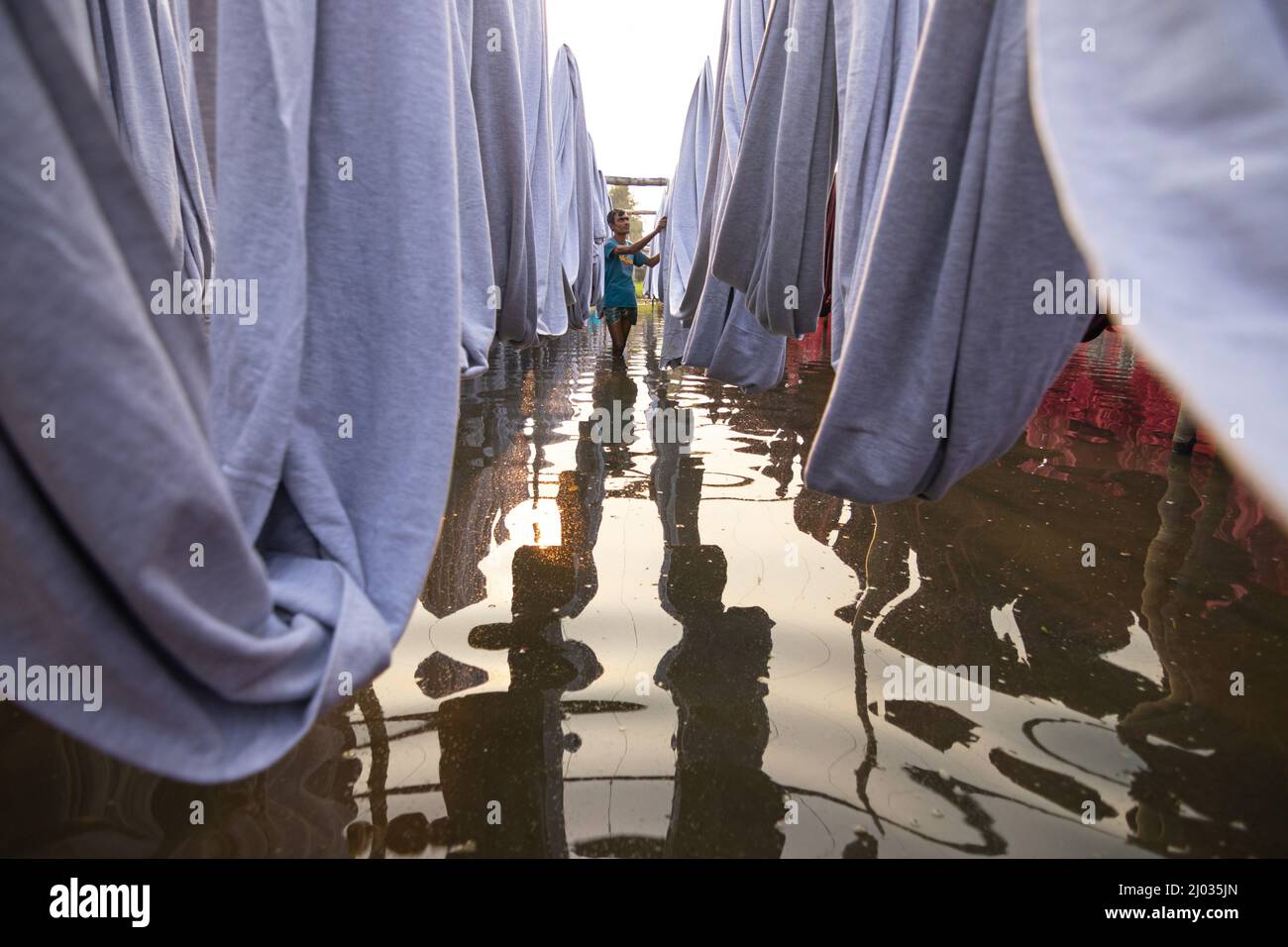 Narayanganj, Dhaka, Bangladesh. 16th Mar 2022. I lavoratori appendono migliaia di diversi tessuti colorati su fili di ferro legati tra un telaio di bambù e costantemente li trasformano in modo che si asciughino perfettamente in campo allagato a Narayanganj, Bangladesh. I fili di ferro sono usati fra una struttura di bambù per generare le linee giganti di lavaggio per la parte finale del processo di tintura mentre i tessuti sono asciugati nel sole. I fili luminosi di panni blu, rosa, arancione e verde-tinti appendono sopra il campo erboso in una rete abbagliante di colori interbloccanti. Questa è la parte finale del processo di tintura dopo la quale il panno è Foto Stock