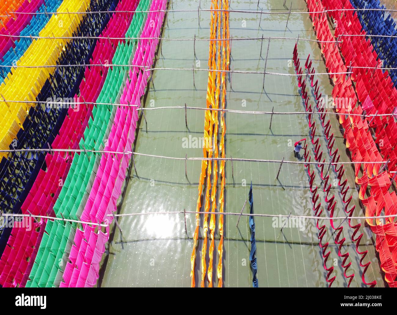 Narayanganj, Dhaka, Bangladesh. 16th Mar 2022. I lavoratori appendono migliaia di diversi tessuti colorati su fili di ferro legati tra un telaio di bambù e costantemente li trasformano in modo che si asciughino perfettamente in campo allagato a Narayanganj, Bangladesh. I fili di ferro sono usati fra una struttura di bambù per generare le linee giganti di lavaggio per la parte finale del processo di tintura mentre i tessuti sono asciugati nel sole. I fili luminosi di panni blu, rosa, arancione e verde-tinti appendono sopra il campo erboso in una rete abbagliante di colori interbloccanti. Questa è la parte finale del processo di tintura dopo la quale il panno è Foto Stock