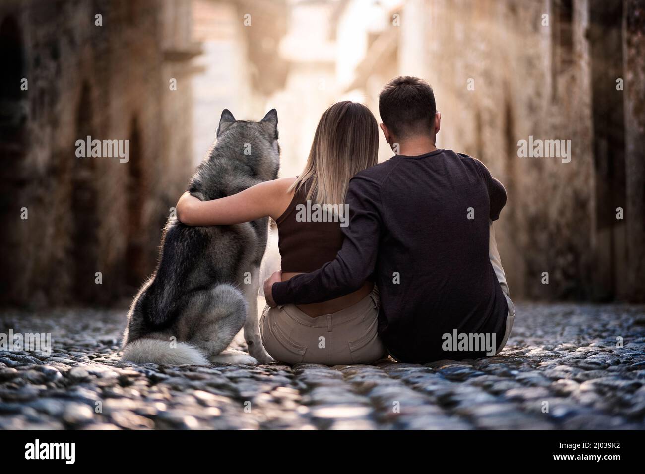 Vista posteriore di due giovani e del loro amato cane lupo seduto e abbracciato in una strada della città vecchia fissando il sole, Piemonte, Italia, Europa Foto Stock