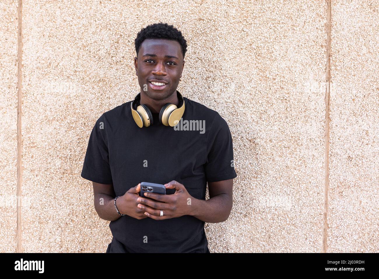 Sicuro di sé positivo giovane africano americano maschile millennial con capelli ricci scuro in t shirt nera e cuffie sul collo sorridente e guardando camer Foto Stock