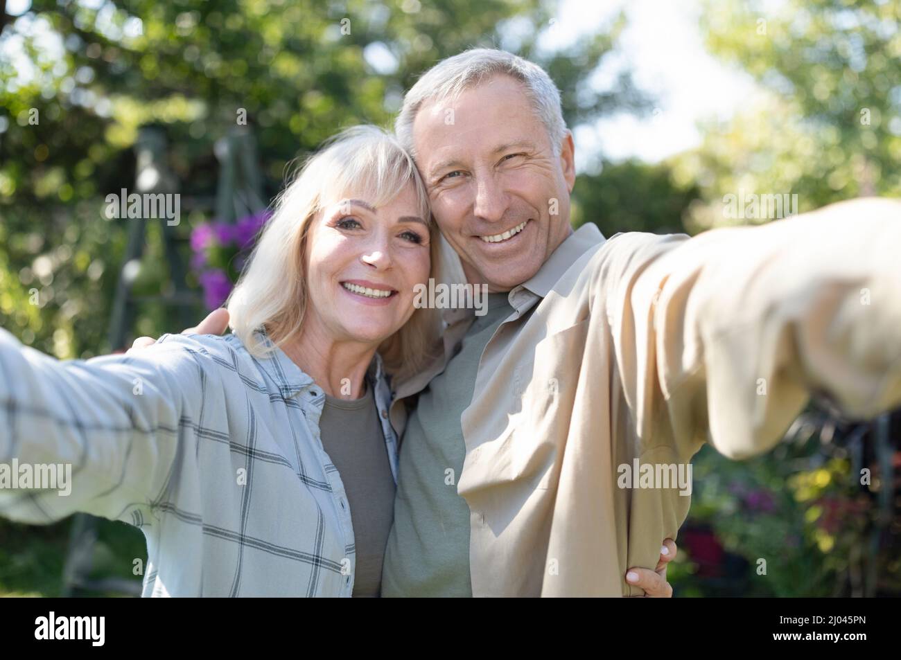 Sposi anziani felici che fanno selfie, abbracciando, sorridendo alla macchina fotografica, trascorrendo il tempo insieme nel loro giardino all'aperto Foto Stock