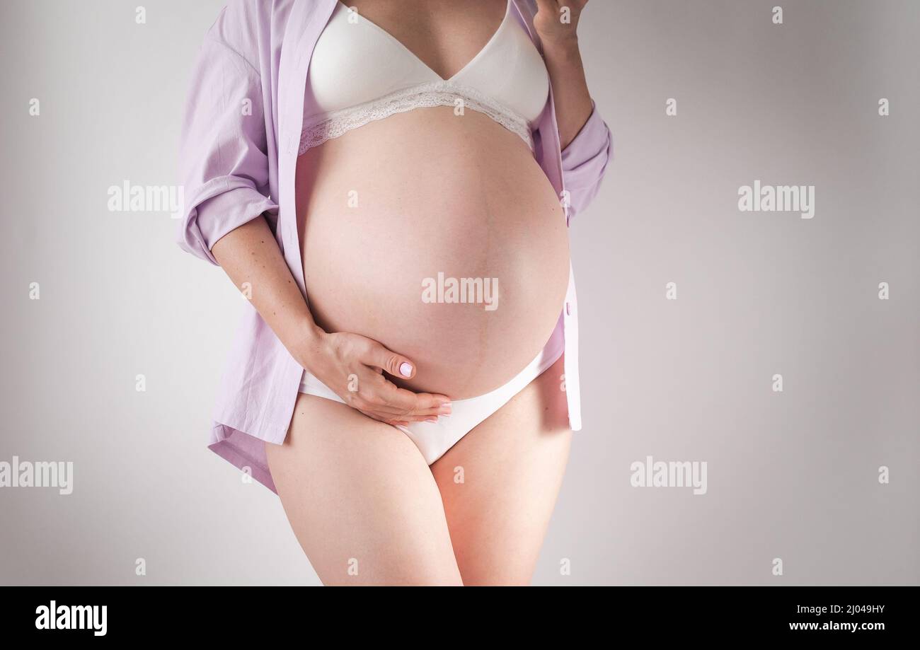 una donna incinta terzo trimestre in intimo e una camicia rosa si erge su uno sfondo bianco Foto Stock
