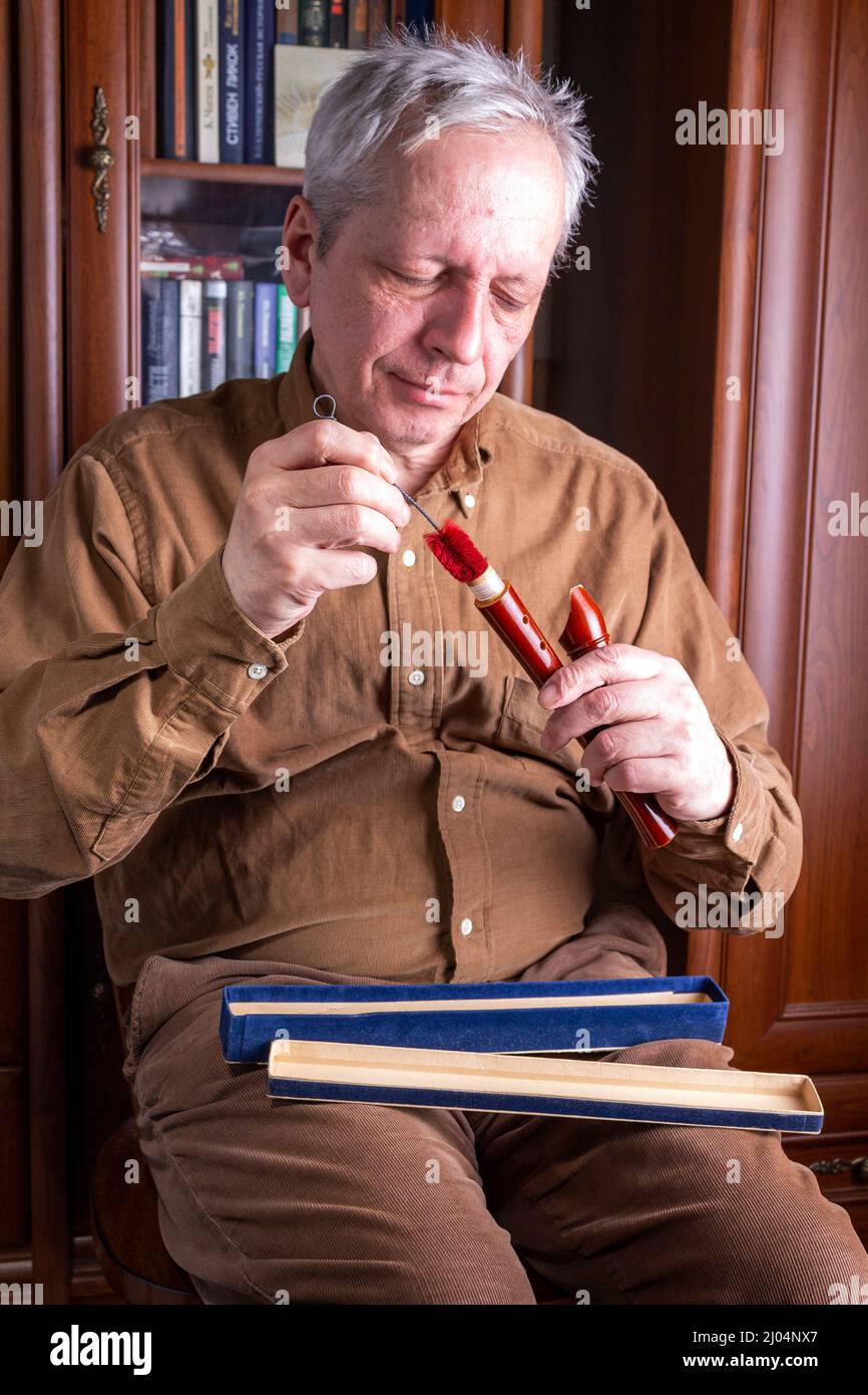 un uomo anziano pulisce un flauto di legno mentre si siede su una sedia dentro Foto Stock