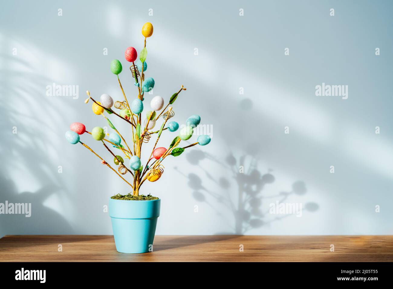 Decorazioni dell'albero di Pasqua in vaso blu con uova colorate sulla console di legno con sfondo chiaro e ombra. Decorazione casa vacanza di Pasqua. Minimalismo, s Foto Stock