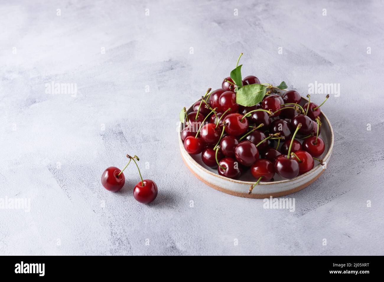 Bacche di ciliegie fresche in un piatto di ceramica leggera su sfondo grigio di cemento. Posiziona per testo. Foto Stock