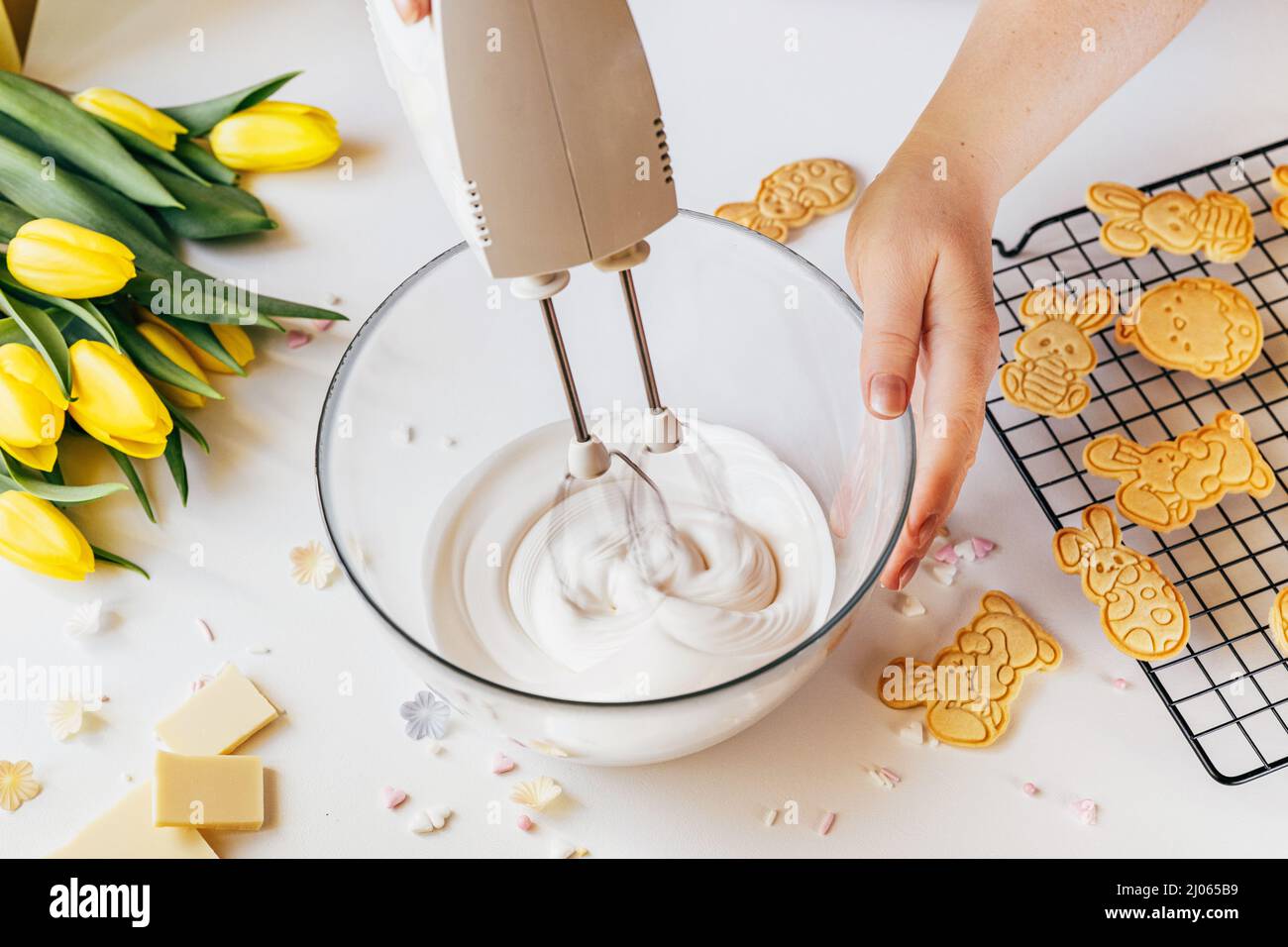 Scatto corto della donna usando la macchina del miscelatore che fa la glassa del biscotto per la decorazione. Donna che tiene un recipiente in vetro con glassa di zucchero e mixer elettrico. Vacanze di Pasqua sostegno Foto Stock