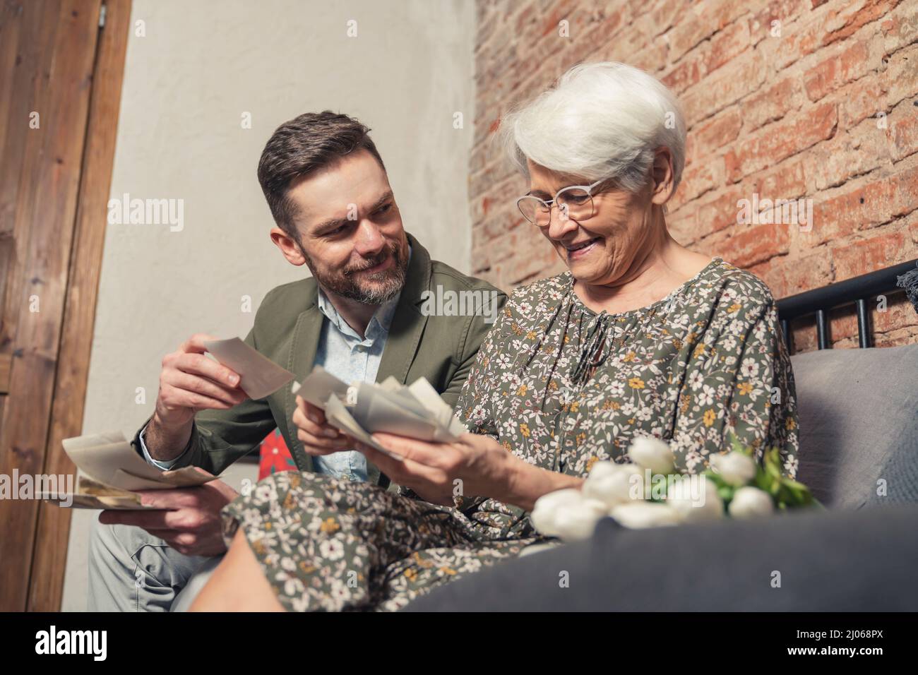 Vivace vecchia donna grigio-capelli pensa volentieri al passato e mostra il suo nipote adulto vecchie fotografie il giorno del nonno. Foto di alta qualità Foto Stock