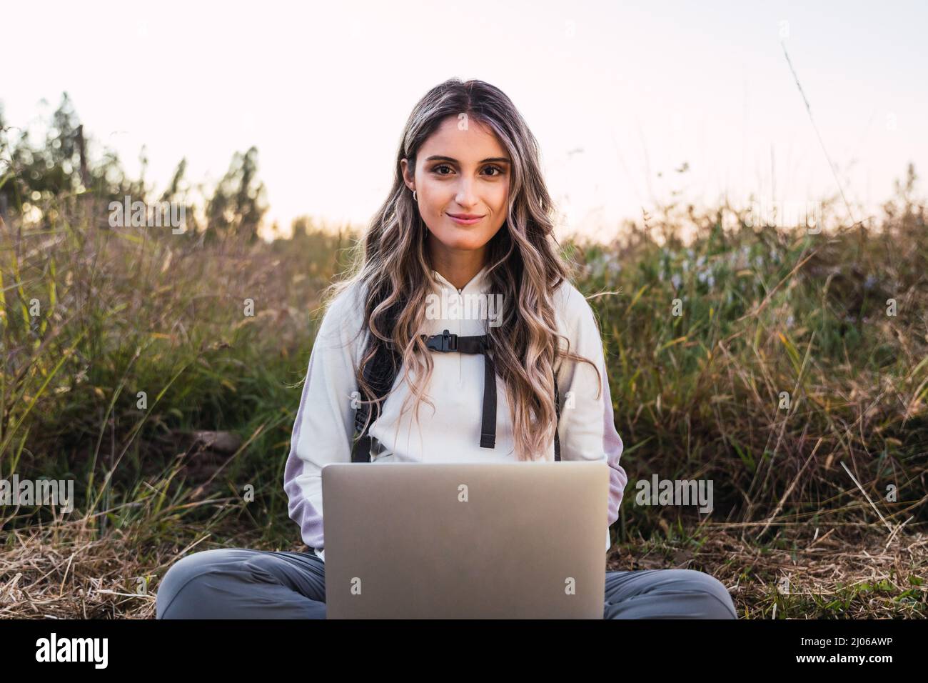 Giovane donna caucasica con uno zaino su, utilizzando un portatile per telelavoro in mezzo al campo. Nomade digitale. Foto Stock