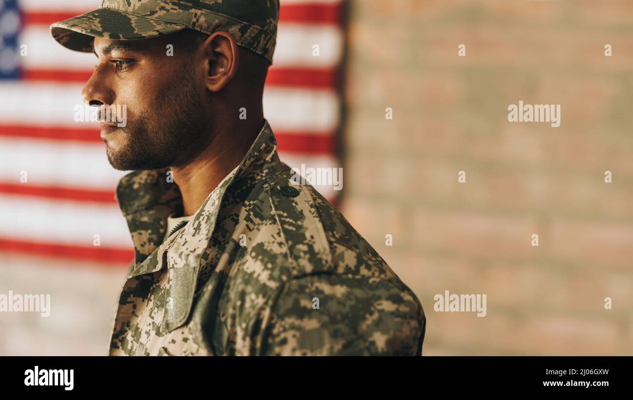 Coraggioso giovane soldato che guarda via con la bandiera americana sullo sfondo. Membro di servizio patriottico che indossa l'uniforme mimetizzata degli Uniti Foto Stock