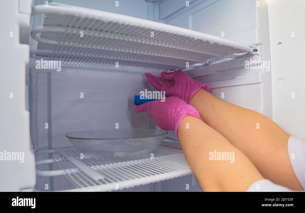 Sbrinamento e pulizia del congelatore. Una donna in lattice guanti fazzoletti meltwater con una spugna. Profondità di campo poco profonda. Foto Stock