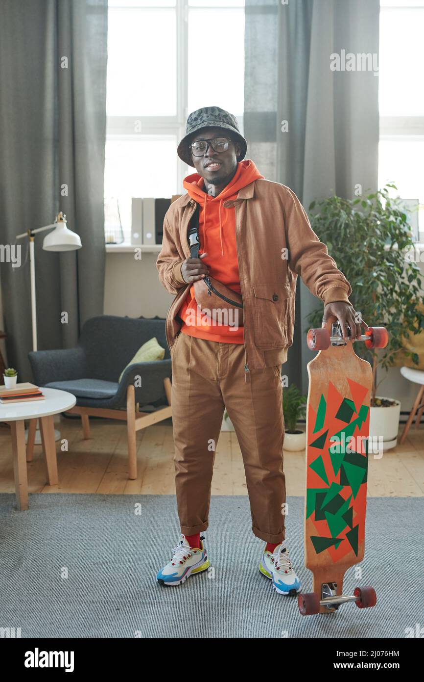 Ritratto verticale a scatto completo di un giovane elegante in piedi in salotto con skateboard pronto per uscire sorridendo alla fotocamera Foto Stock