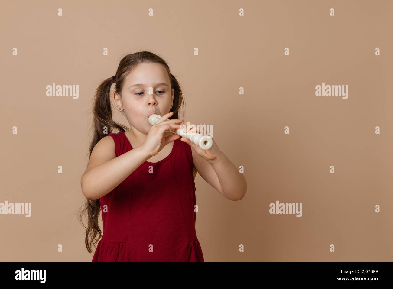 Ragazza in abito rosso sedulente giocare melodia sul flauto, soffiare aria nel condotto, sfondo beige. Imparare a suonare lo strumento musicale woodwind. Foto Stock