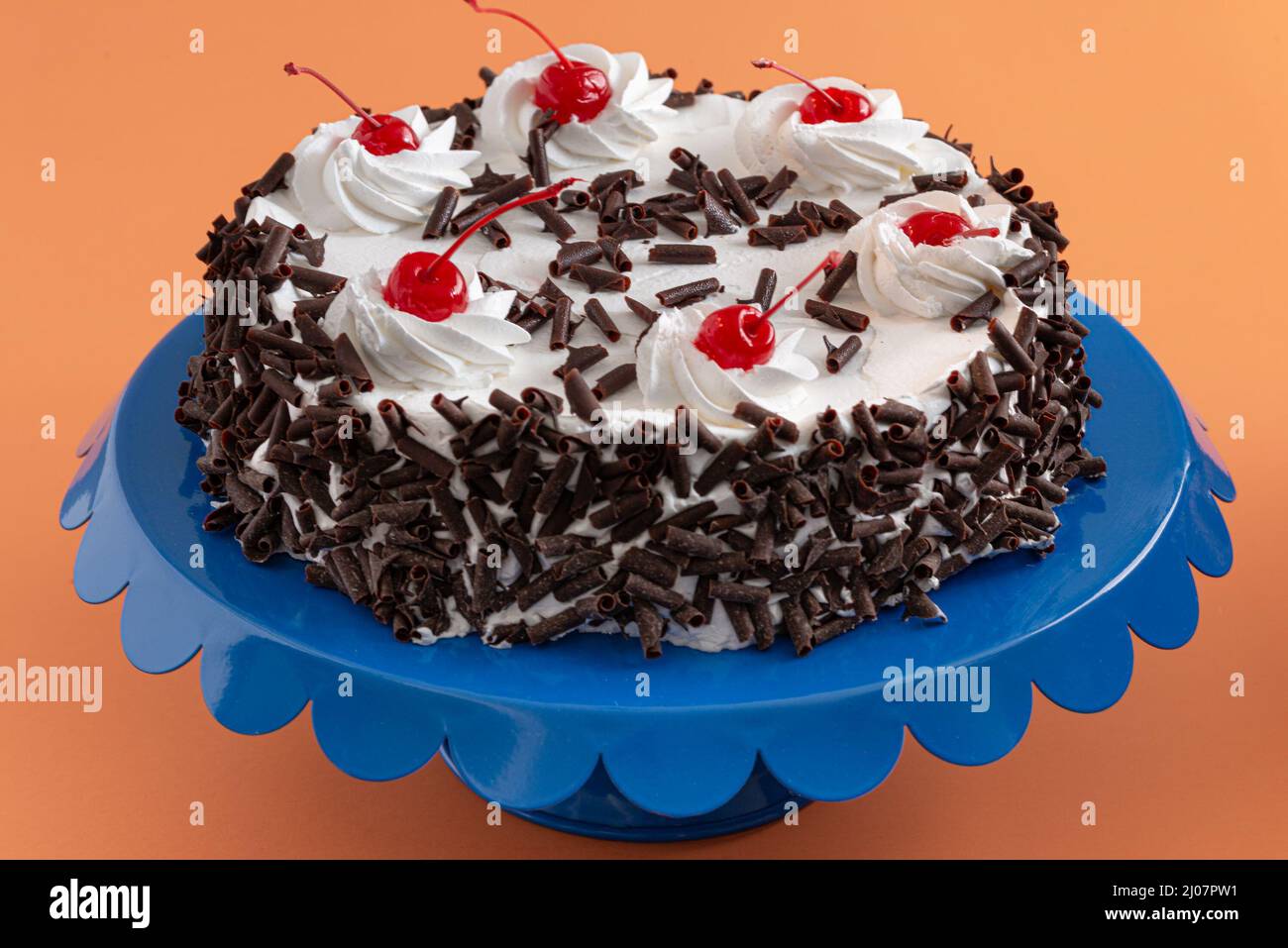 Una torta lussuosa ricoperta di panna montata e scaglie di cioccolato, condita da ciliegie rosse brillanti. Posizionato su un vivace supporto per torte blu che contrasta Foto Stock