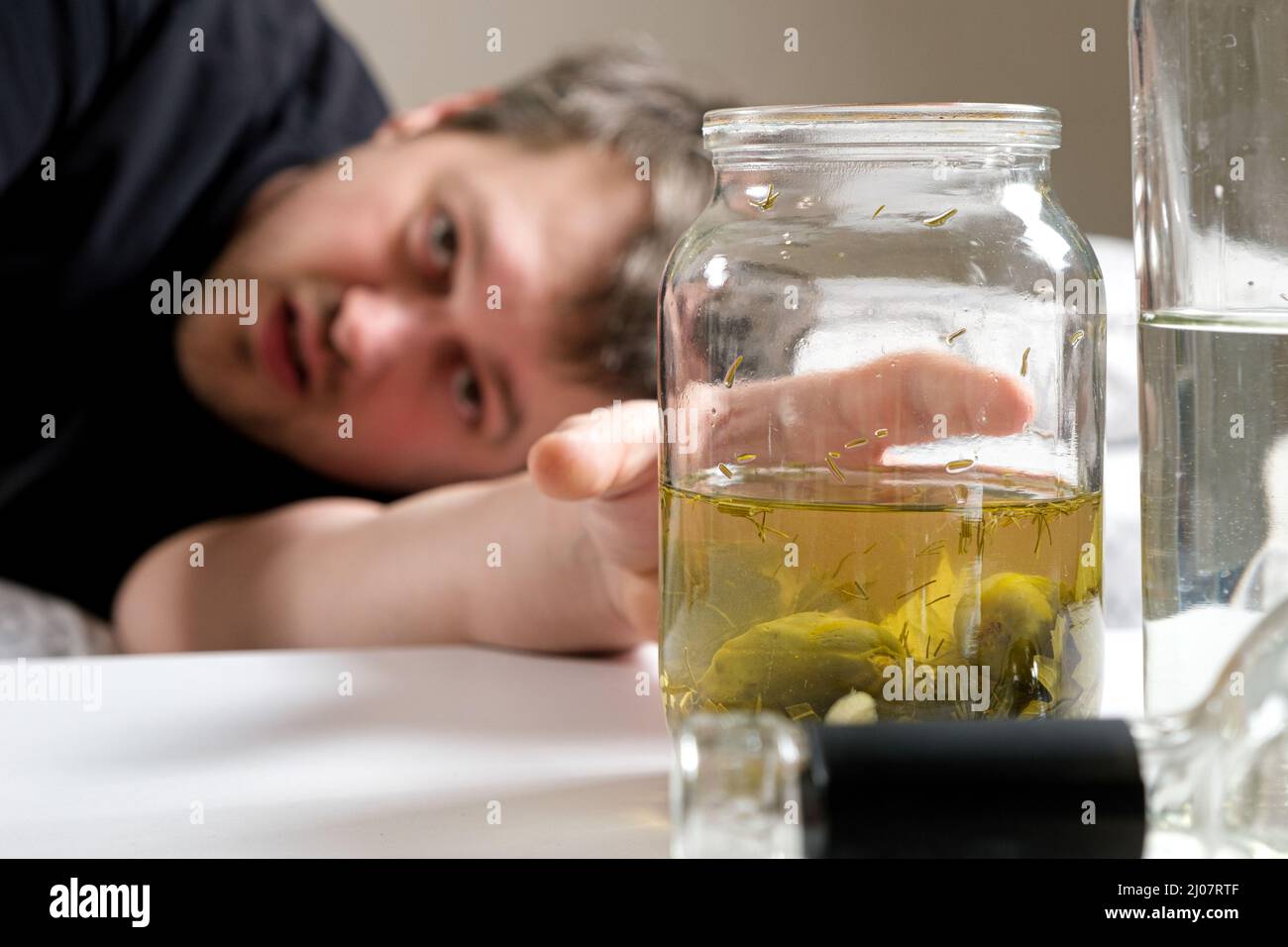 Un uomo con una dipendenza da alcol si trova sul letto e raggiunge con la mano un vaso di cetriolo salato salamoia. Rimedi popolari per i hangovers Foto Stock