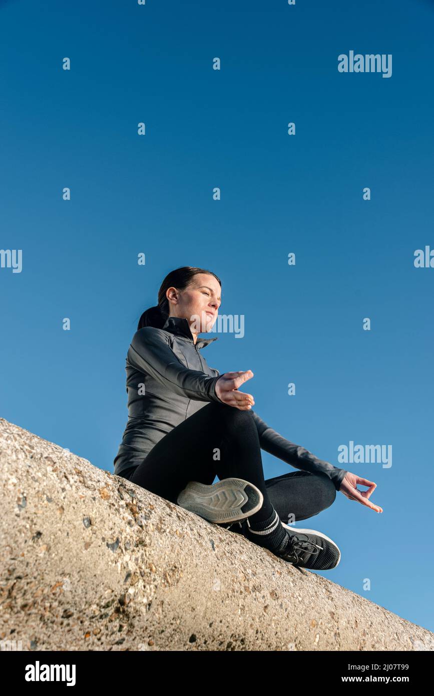 Donna seduta a gambe incrociate, pratica yoga e meditazione all'aperto con un cielo blu. Foto Stock