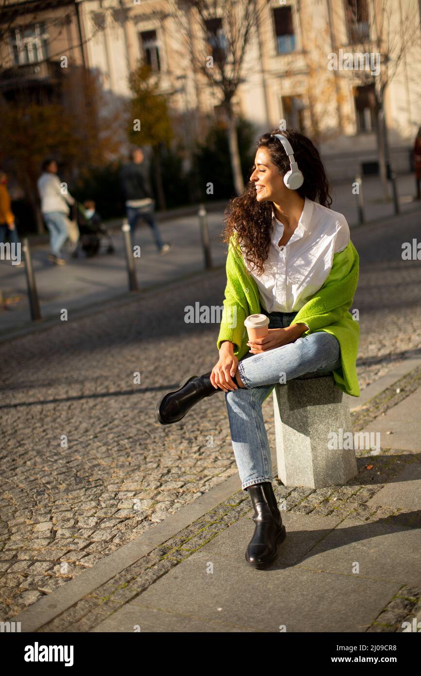 Bella giovane donna che ascolta musica con smartphone in strada e tiene il caffè da asporto Foto Stock