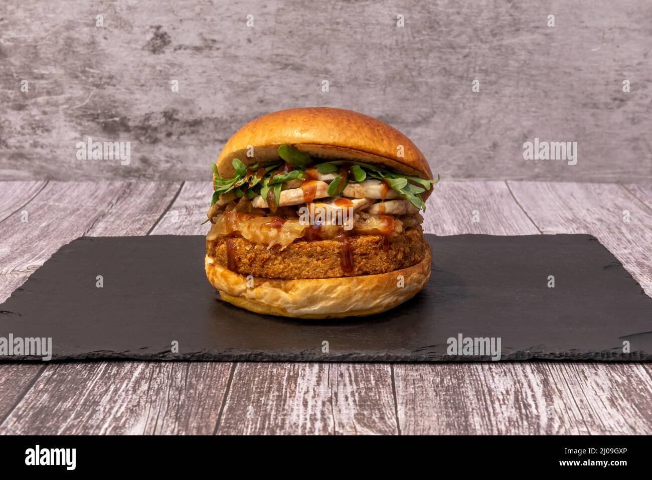 Hamburger di bistecca vegano con funghi, salsa barbecue, foglie di rucola fresche e cipolle caramellate su un piatto di ardesia nera Foto Stock