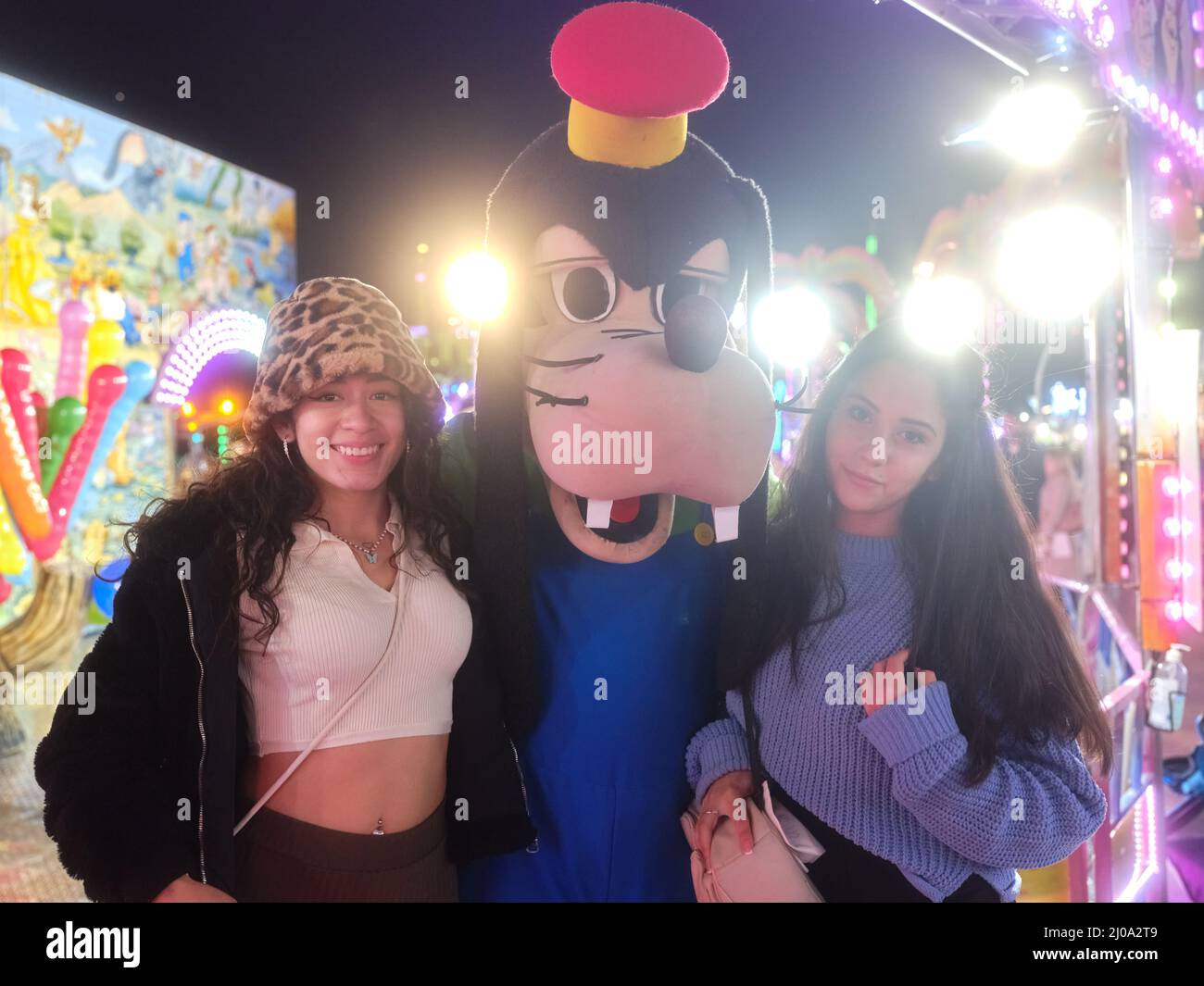 Due donne latina in piedi accanto ad un attore vestirsi come cane in una fiera di notte Foto Stock