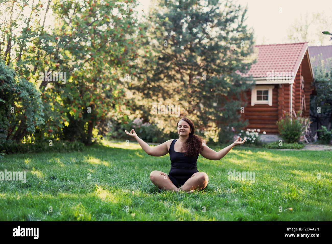 Giovane donna sovra-pesata che fa yoga seduto in posizione di loto fuori sul cortile di cottage con alberi e casa di legno in background. Corpo positivo Foto Stock