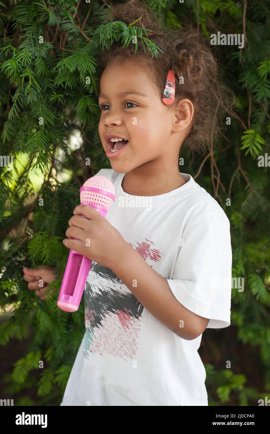 Simpatica piccola cantante ragazza che canta sul microfono nel parco della città circondato da vegetazione verde Foto Stock