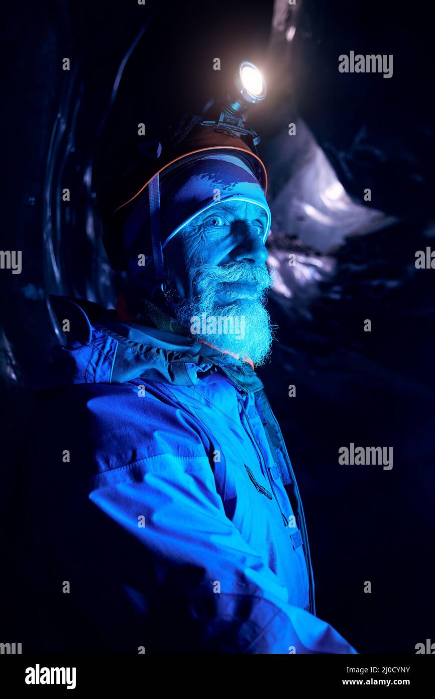 Vecchio alpinisto con barba grigia in casco con lampada a incandescenza in una profonda grotta di ghiaccio glaciale scuro con luci blu esplorare il ghiacciaio invernale di montagna in Kazakhstan Foto Stock