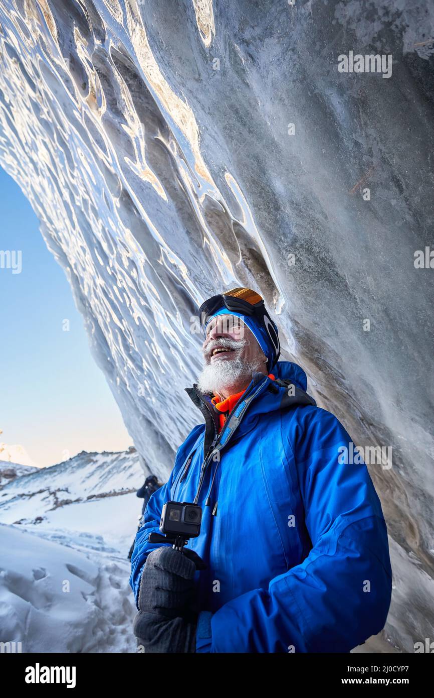 Ritratto di vecchio uomo bearded con macchina fotografica di azione in costume blu vicino parete della grotta di ghiaccio al ghiacciaio nella valle di montagna coperta di neve in Almaty, Kazakhstan Foto Stock