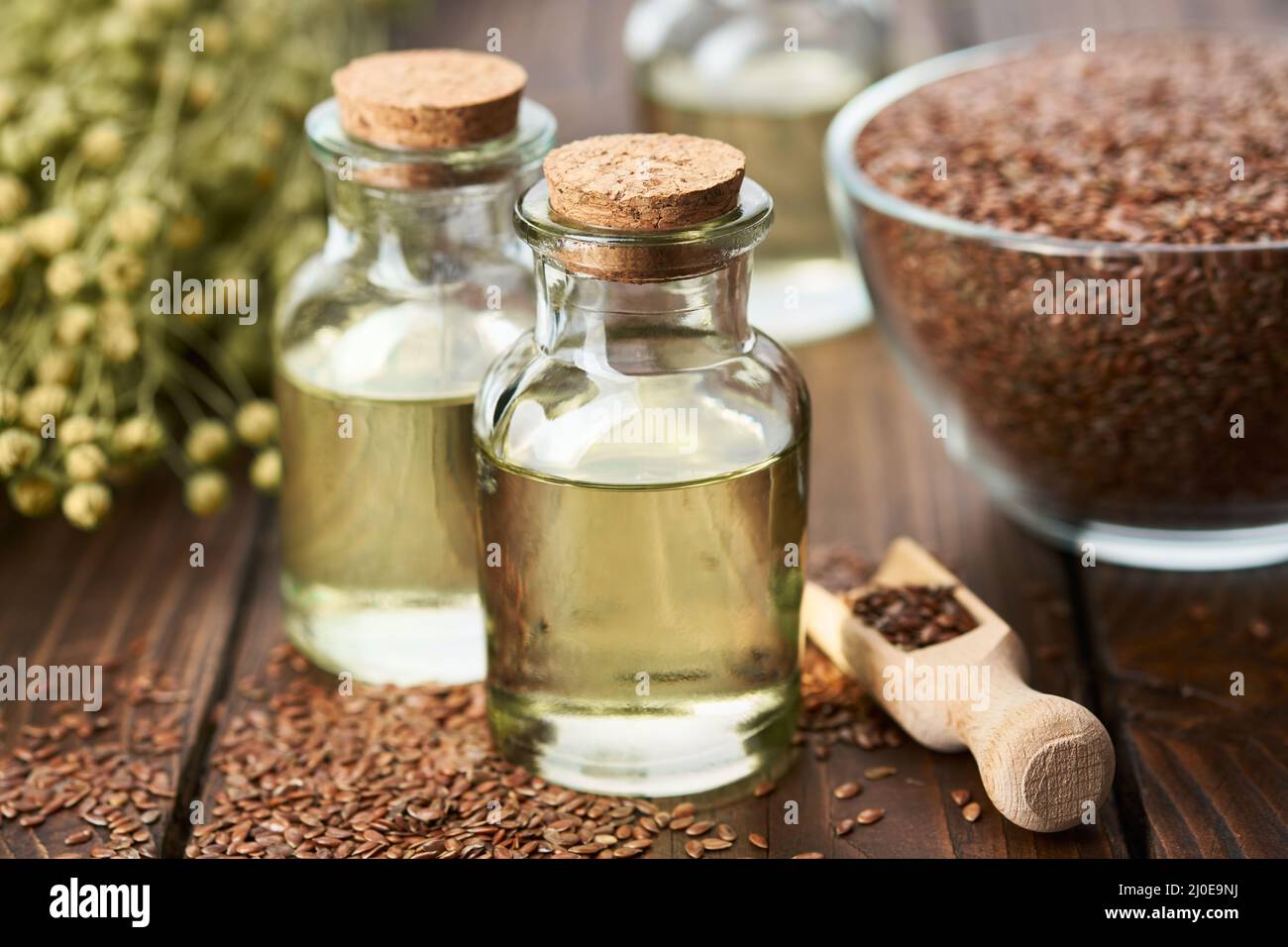 Due bottiglie di olio di lino fresco e ciotola di vetro di semi interi di lino. Fascio di piante di lino su tavola. Foto Stock