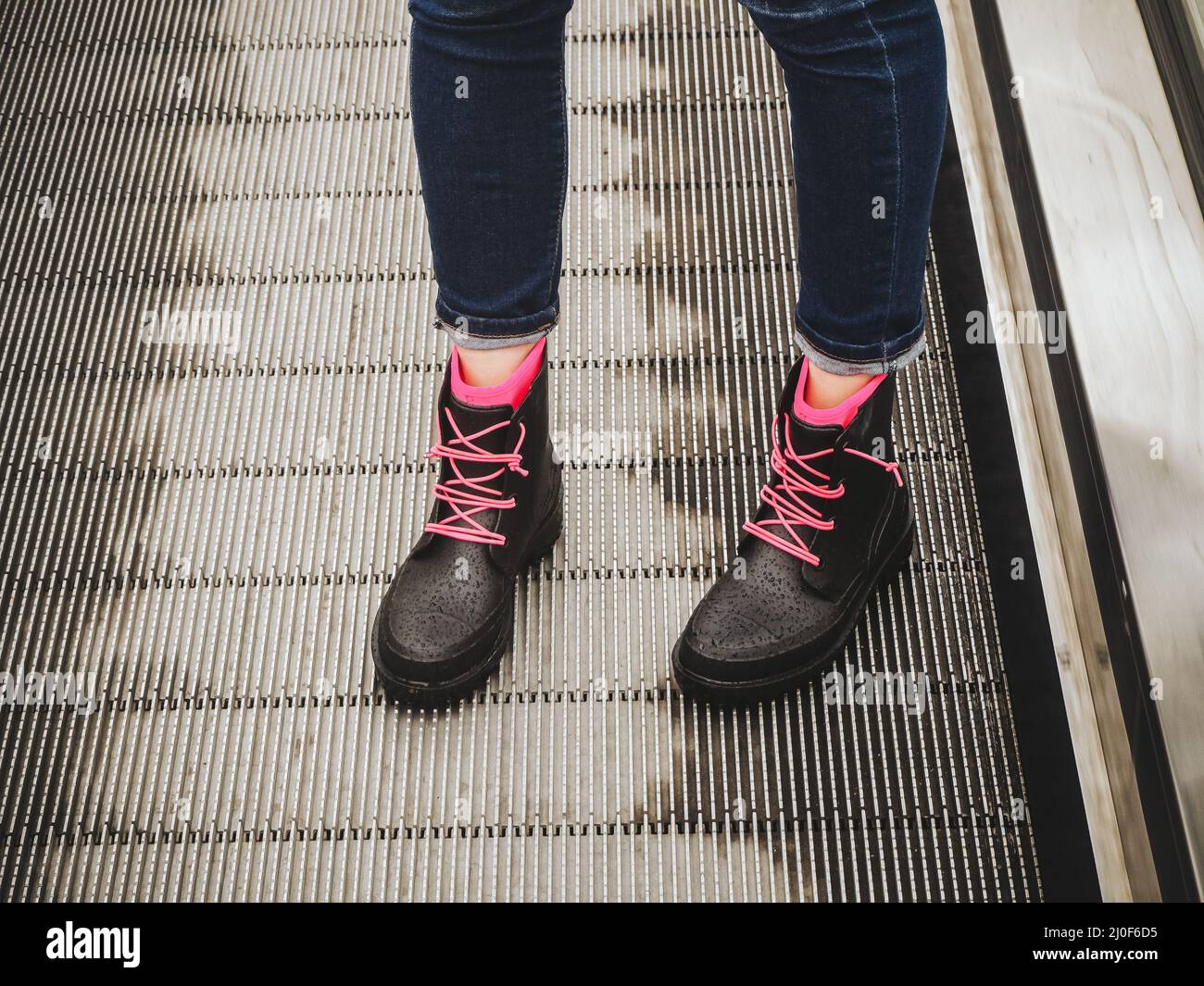 Le gambe femminili in jeans blu e gli stivali in gomma nera bagnata con lacci rosa si trovano sulla scala mobile Foto Stock
