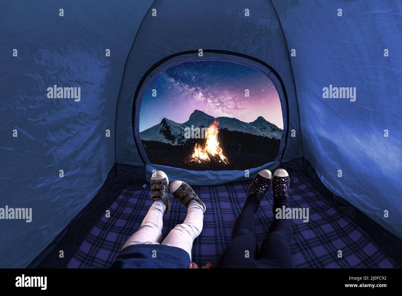Due bambini all'interno su una tenda da campeggio che guarda stelle e montagne di notte con un fuoco acceso Foto Stock