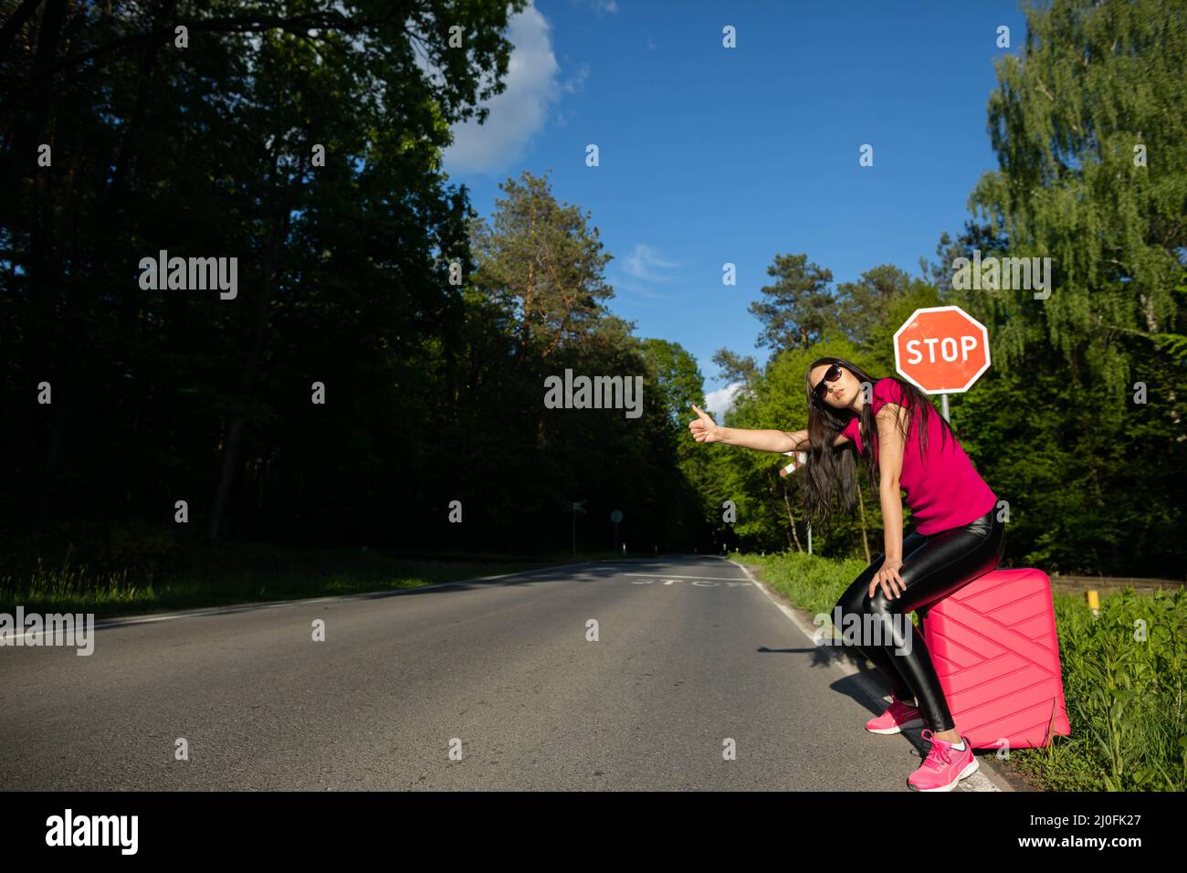 Un giovane adulto si erge sul lato della strada con una valigia rosa e cerca di fermare le altre auto per ottenere un giro gratuito per il hol Foto Stock