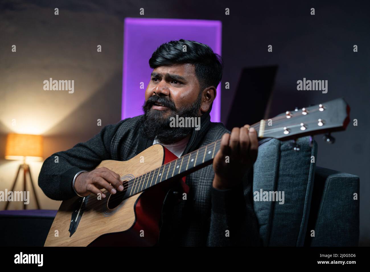 Giovane beardman che suona la chitarra mentre si siede sul divano a casa - concetto di musicista, artista e hobby. Foto Stock