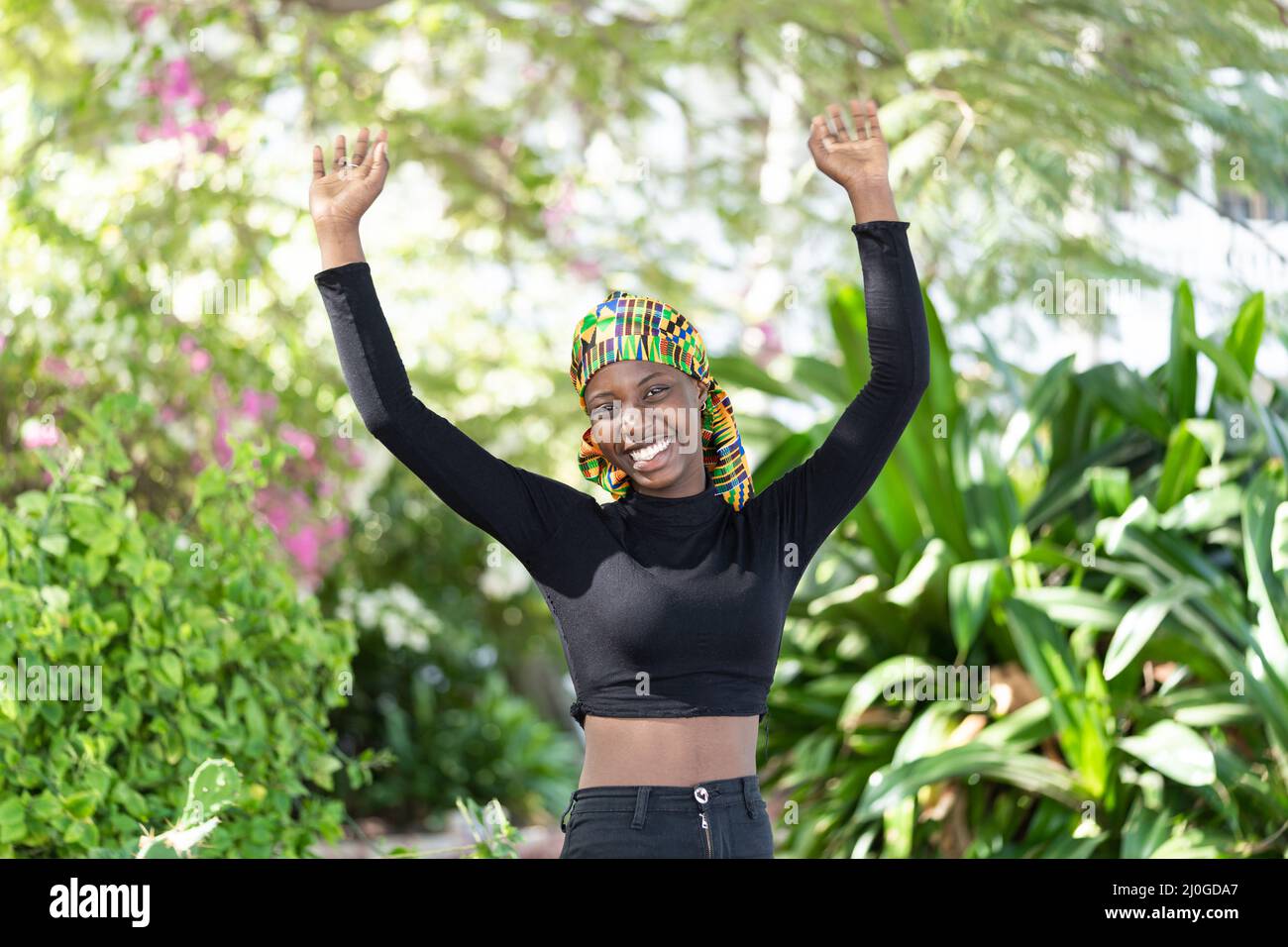 Splendida ragazza africana in pantaloni e Long-sleeved nero crop top in piedi in un giardino fiorente con un grande sorriso, sollevando le braccia al cielo come un Foto Stock