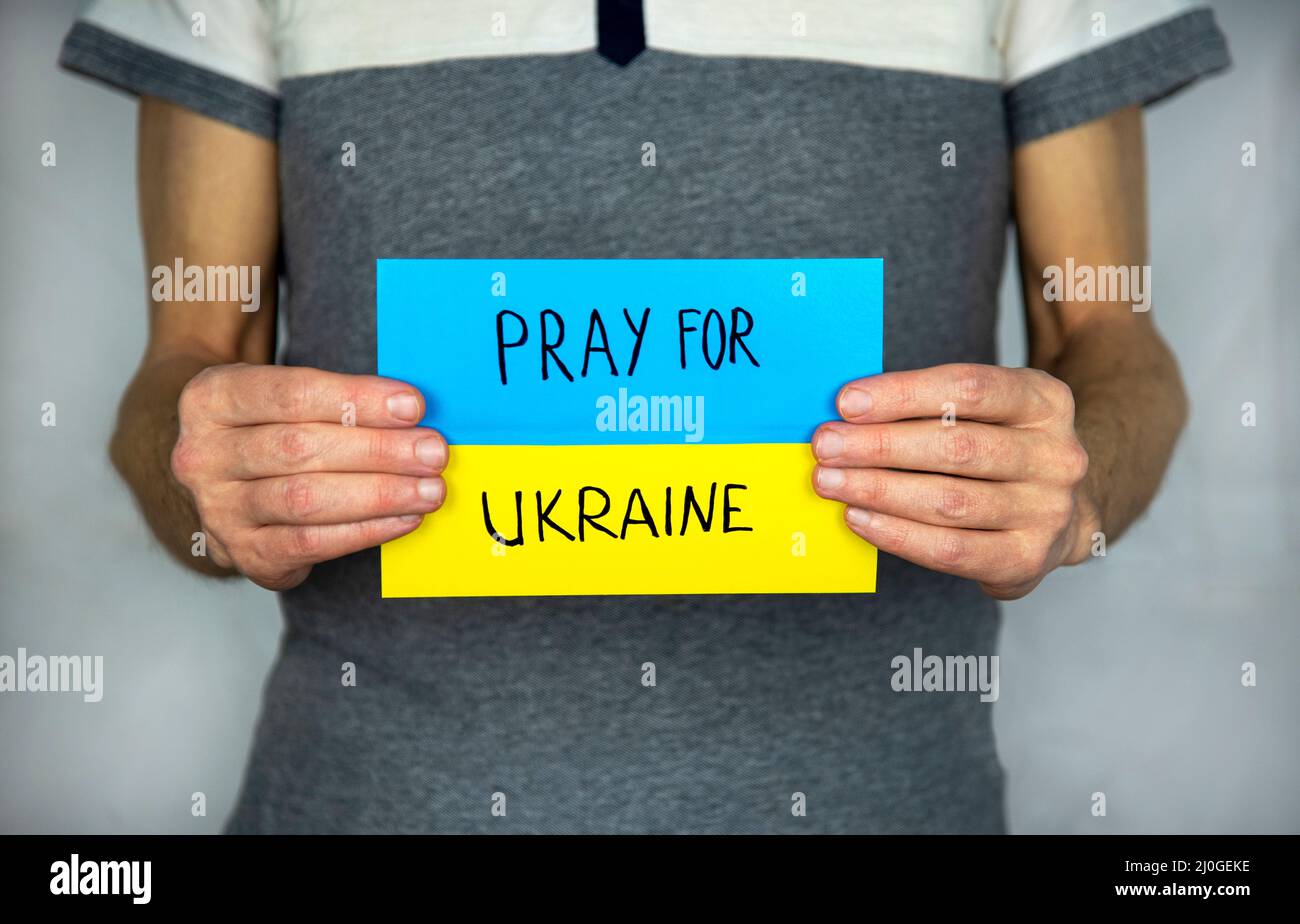 Un uomo in piedi su un grigio, tiene in mano lo sfondo di un cartoncino giallo-blu bandiera Ucraina con l'iscrizione "prega per l'Ucraina" Foto Stock