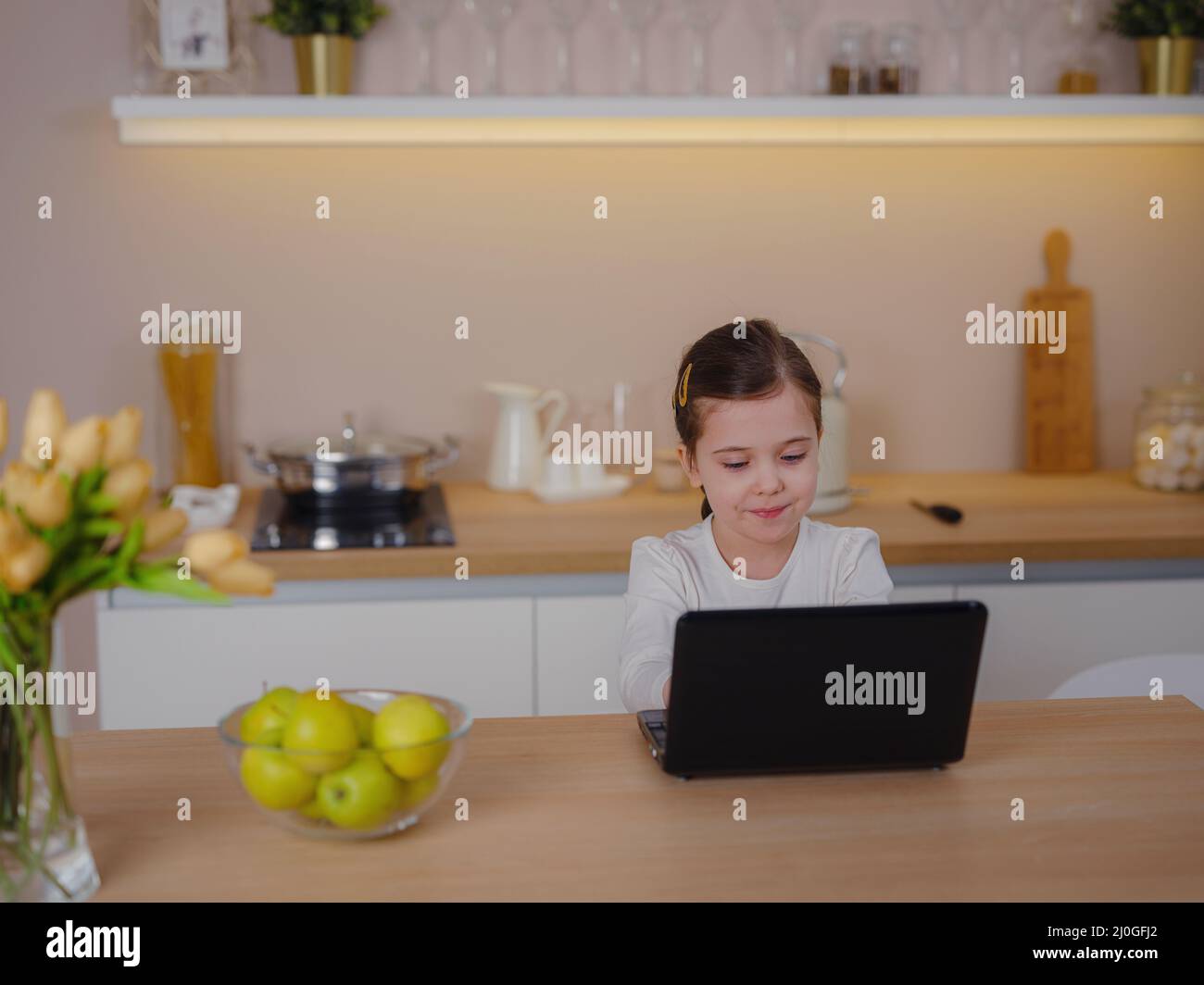 piccola ragazza caucasica ha videochiamata in classe remota con l'insegnante che usa il laptop, imparare online sul computer, concetto di apprendimento a casa. bambino codifica di fronte al portatile in cucina Foto Stock